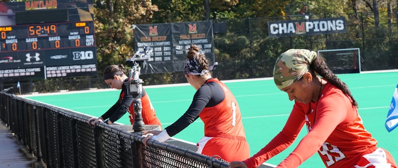 A group of athletes wearing orange uniforms are on the sideline of a sports field, resting against a fence. Behind them, a large digital scoreboard displays team names and scores. The field is bright green, and several banners with sponsor logos and championship wins are visible. The athletes appear to be taking a break, possibly before or during a field hockey game.